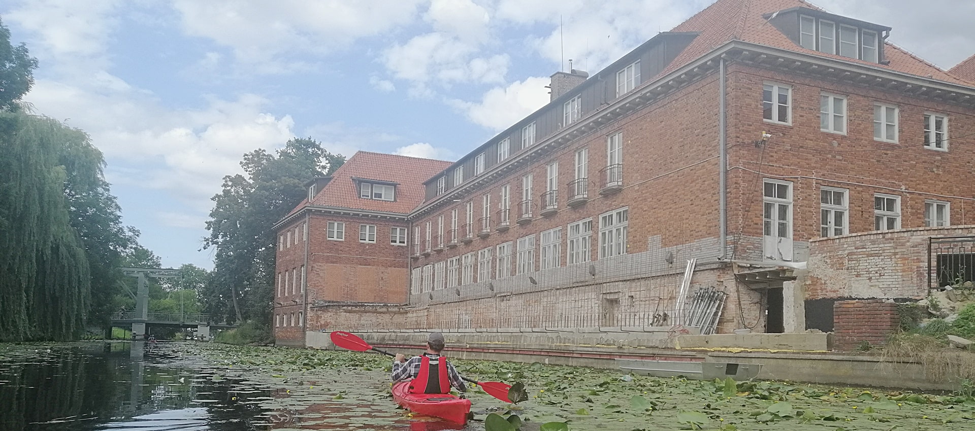 mężczyzna płynący czerwonym kajakiem obok dużego budynku z pomarańczowej cegły