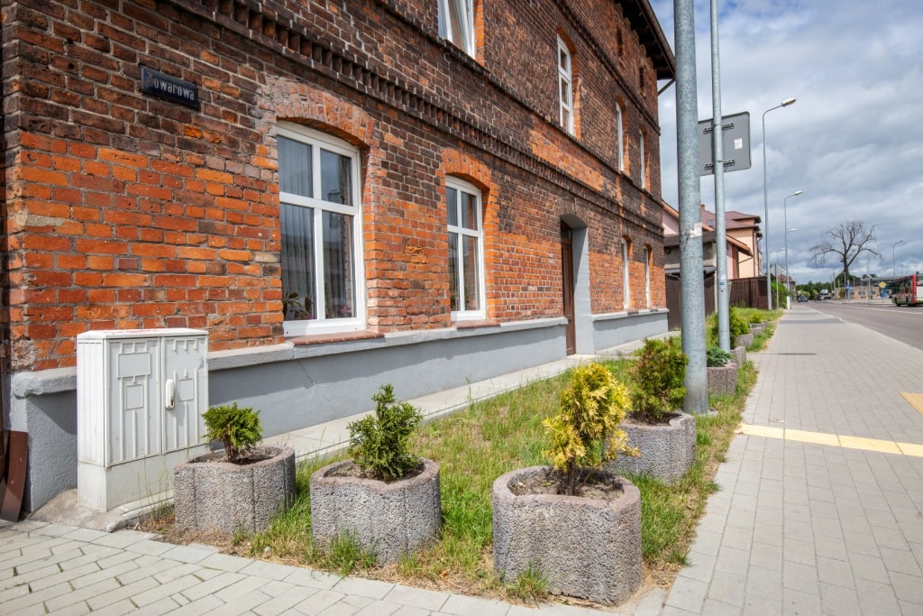 Zdjęcie przedstawia piętrowy budynek z czerwonej cegły oraz chodnik i wąski trawnik z małymi krzakami.