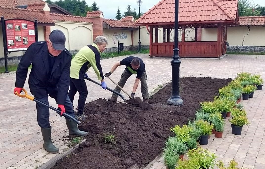 Dwóch mężczyzn i jedna kobieta przekopujący ziemię w celu umieszczenia nasadzeń zieleni w nieutwardzonym fragmencie placu. Plac otacza jasny mur, przy którym stoi drewniana altana z brązowym dachem.