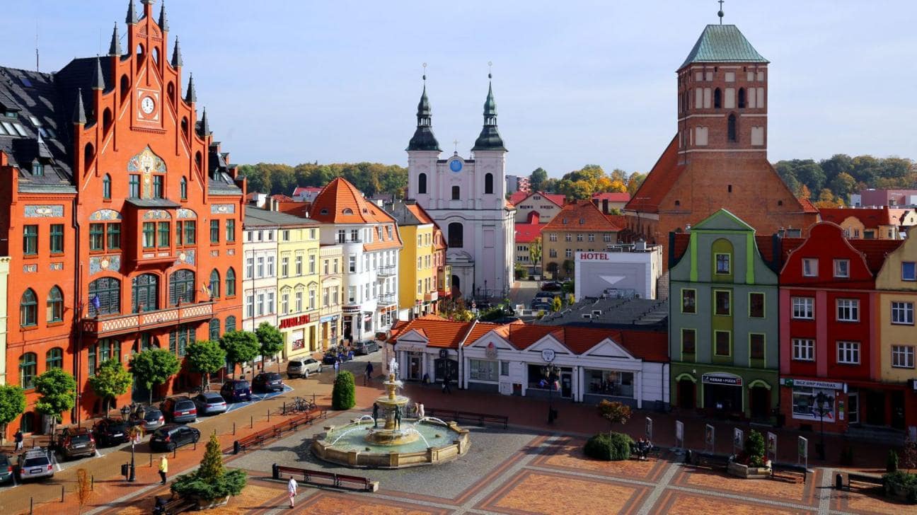 Na zdjęciu widać mały rynek miejski z fontanną na środku i historyczną zabudową z różnokolorową elewacją. Na zdjęciu świeci słońce i jest błękitne niebo.