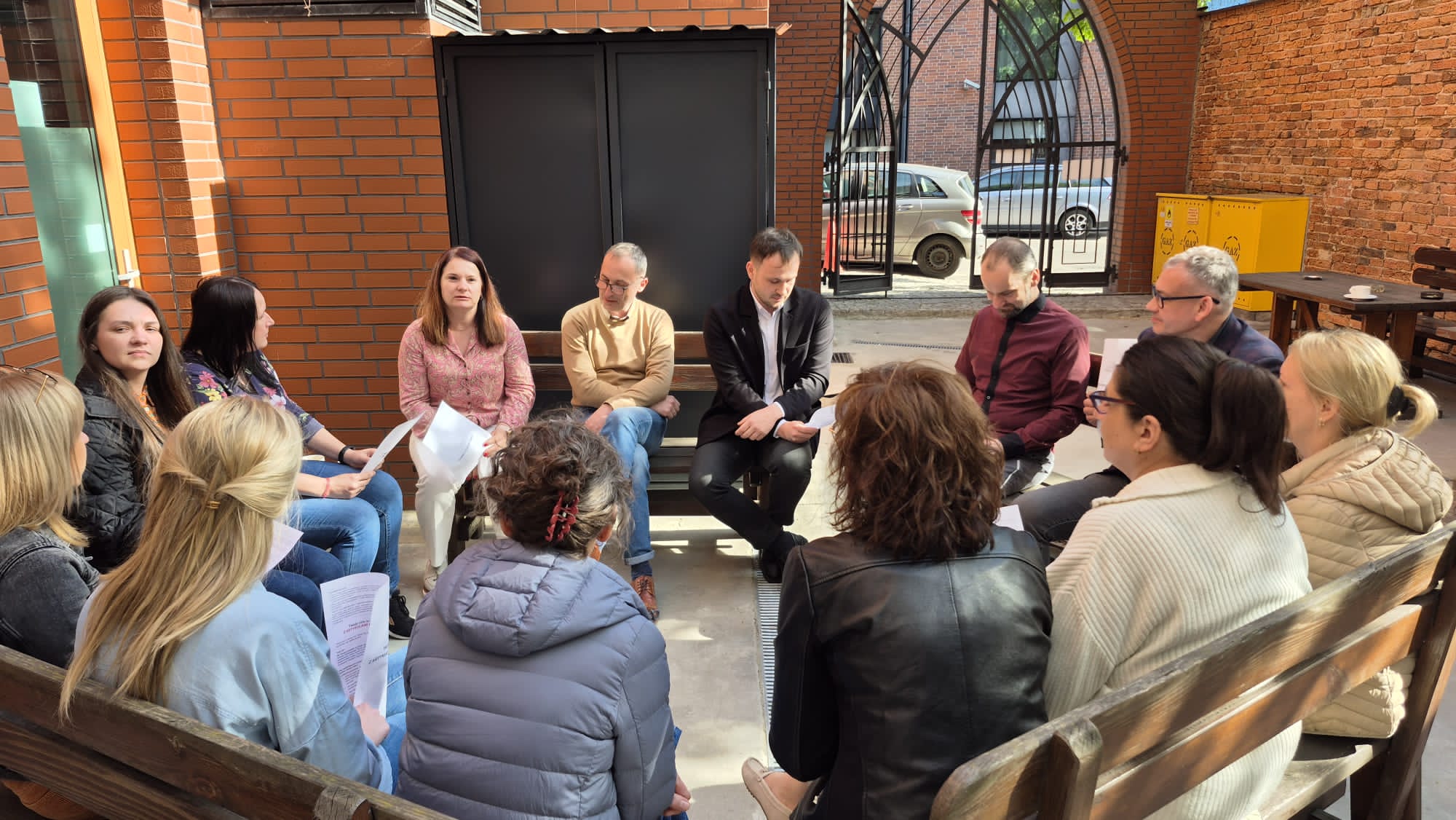 Grupa ludzi siedząca na podwórku na ławkach ułożonych w koło