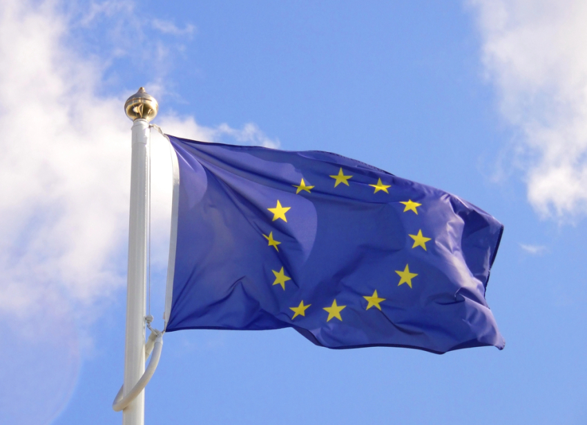 Flaga Unii Europejskiej w kształcie prostokąta powiewa na wietrze. Dwanaście żółtych gwiazd w kręgu na niebieskim tle.