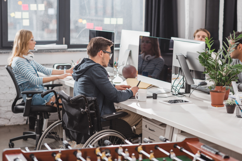 W biurze kilka osób siedzi przy biurkach i pracuje na komputerach. Jednym z pracowników jest mężczyzna na wózku inwalidzkim.