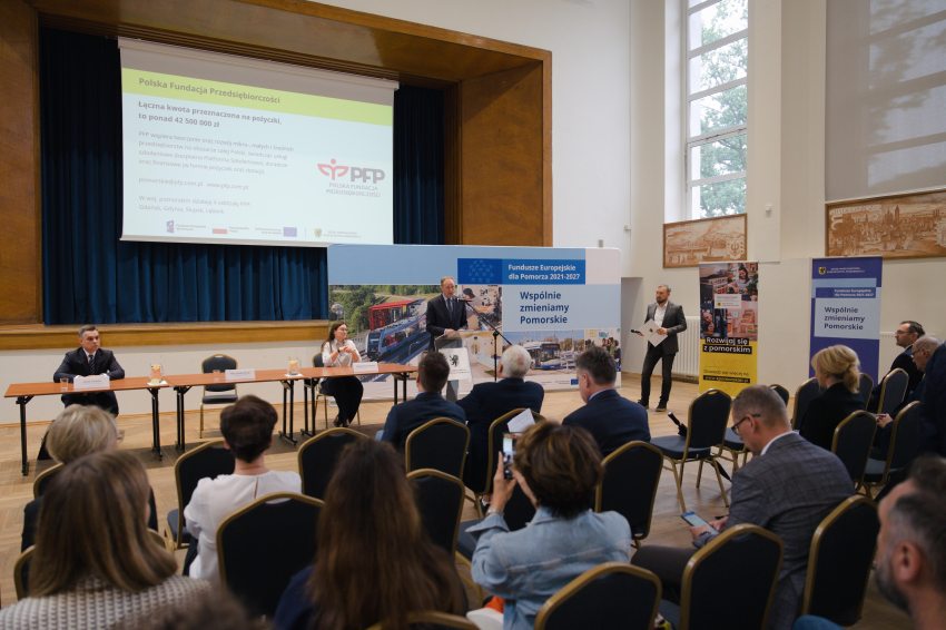 Widok sali. Osoby siedzą na krzesłach. W tle prezentacja