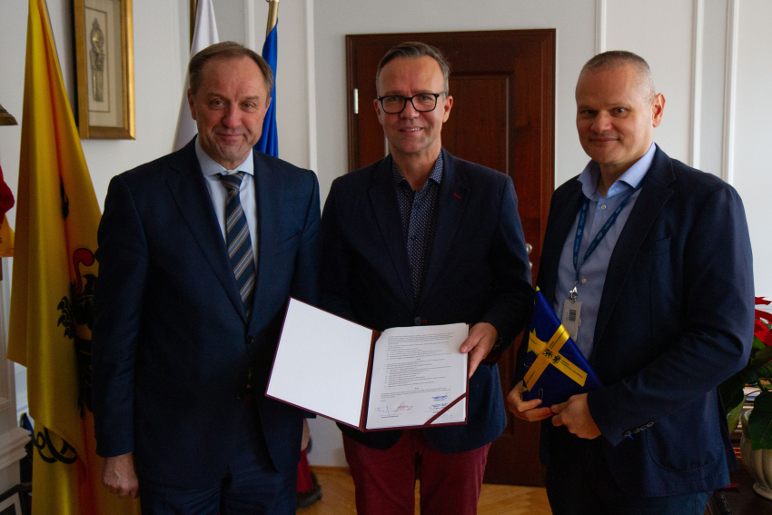 Trzy osoby w gabinecie. Mężczyzna w środku trzyma umowę. Po lewej stronie Marszałek Mieczysław Struk