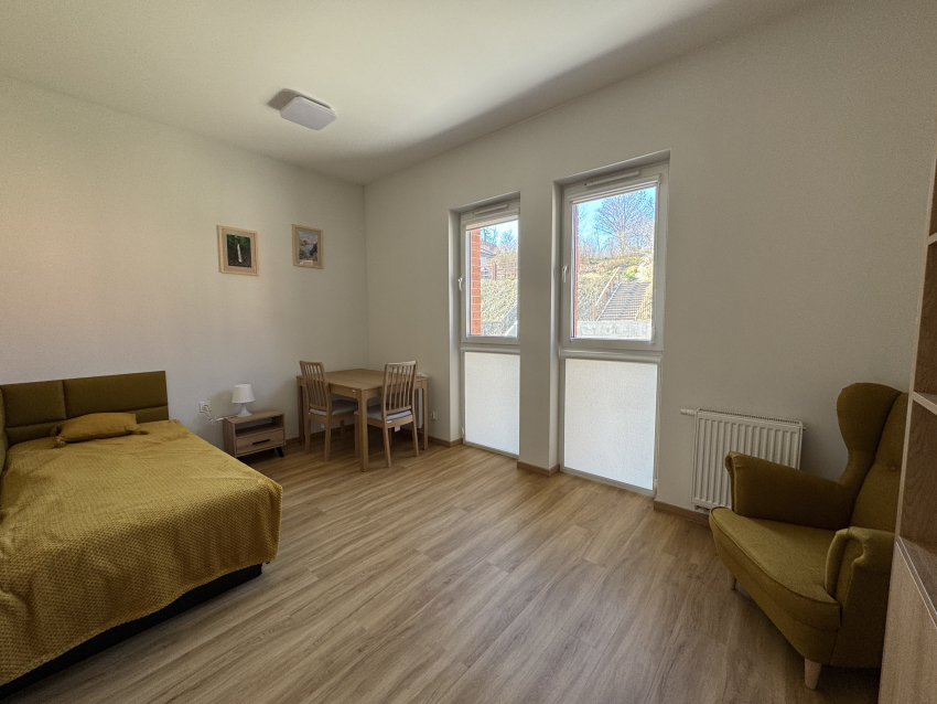 Pokój dla osoby z autyzmem w budynku. W ujęciu łóżko, stół z krzesłami, fotel i dwa okna.