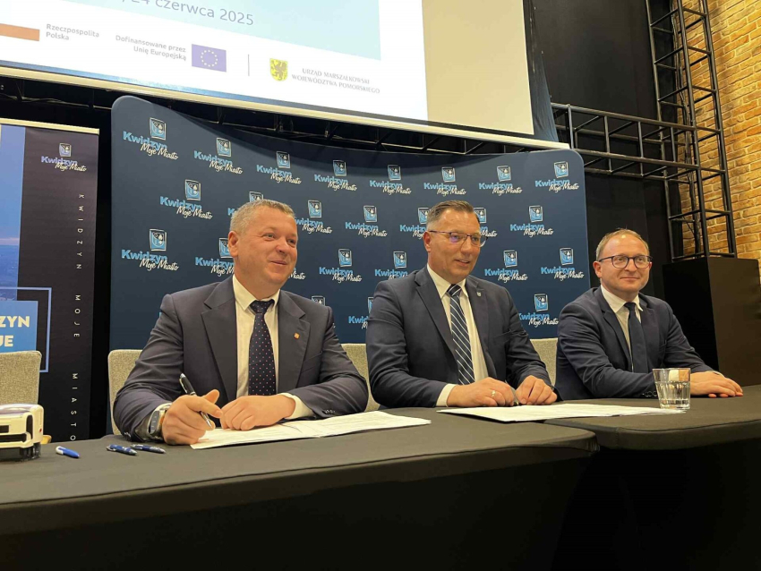 Zdjęcie przedstawia trzech mężczyzn ubranych w garnitury przy stole prezydialnym.