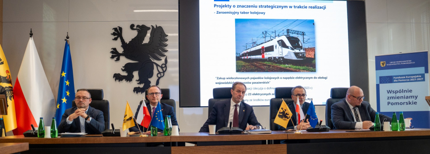 Pięć osób przy stole. W tle flagi i ekran