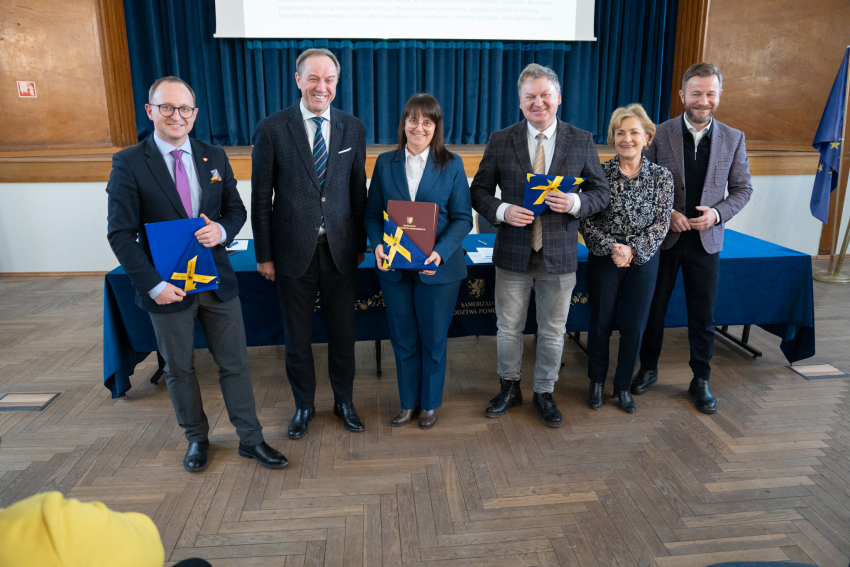 Sześć osób pozuje do zdjęcia prezentując podpisaną umowę oraz flagę Unii Eurpejskiej