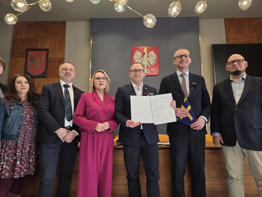 Grupa sześciu osób w strojach wizytowych stoi w rzędzie w oficjalnym pomieszczeniu. Centralna postać, mężczyzna w ciemnym garniturze i okularach, trzyma otwarty, podpisany dokument. Po jego prawej stronie stoi kobieta w wyrazistym, amarantowym garniturze. Na ścianie za nimi widnieje godło Polski (orzeł biały na czerwonej tarczy) oraz herb z lwem. W tle widać nowoczesne oświetlenie sufitowe oraz drewniane panele ścienne. Oficjalne podpisanie porozumienia lub uroczystość urzędową.