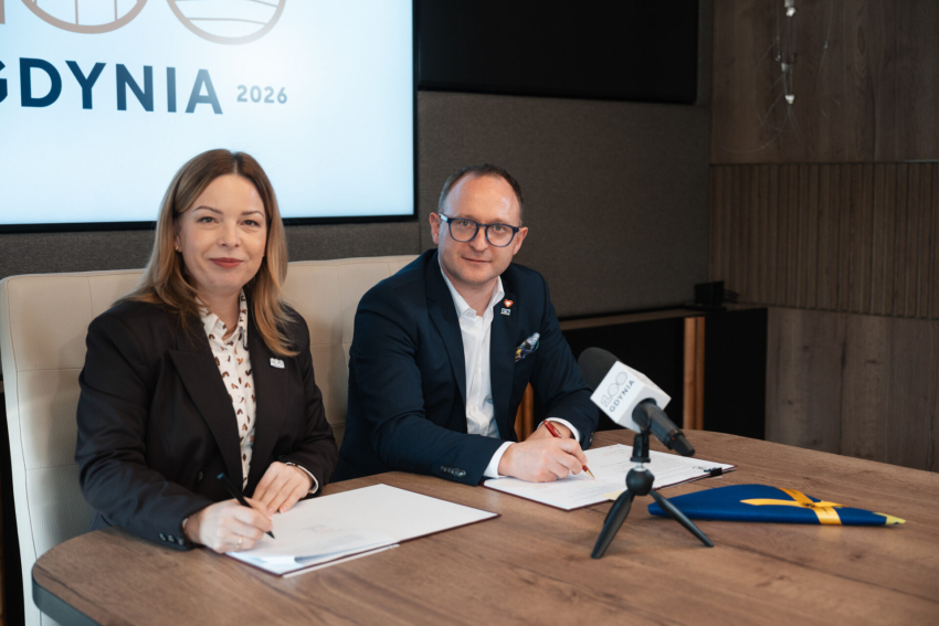 Dwie osoby w strojach biznesowych, kobieta i mężczyzna, siedzą przy drewnianym stole i podpisują dokumenty. Oboje uśmiechają się w stronę obiektywu. W tle widoczny jest duży ekran z napisem „GDYNIA 2026”, a na stole przed nimi stoi mikrofon z logotypem oraz flaga UE