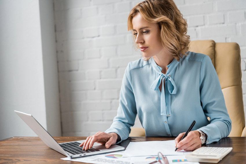 Kobieta trzyma długopis w w dłoni. Drugą ręką pisze na klawiaturze laptopa.