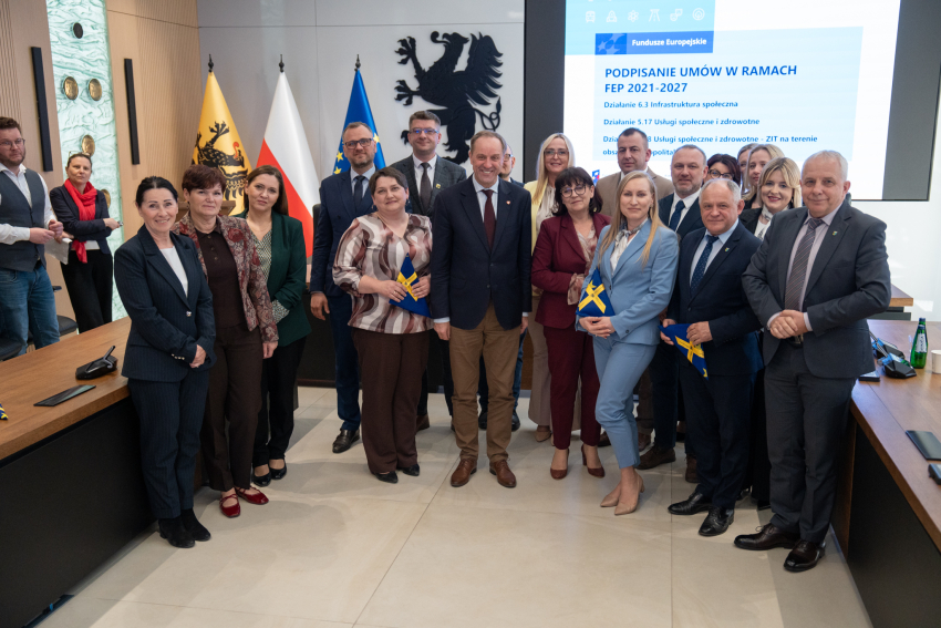 dwadzieścia jeden osób pozuje do zdjęcia prezentując podpisane umowy oraz trzymając flagi unijne. Wśród nich marszałek województwa pomorskiego Mieczysław Struk, oraz przedstawiciele lokalnych władz.