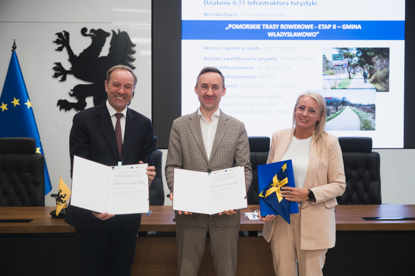 Marszałek Województwa Pomorskiego - Mieczysław Struk, burmistrz Władysławowa - Roman Kużel oraz skarbnik Władysławowa - Kornelia Wittbrodt pozują do uroczystego zdjęcia z podpisaną umową o dofinansowanie i flagą Unii Europejskiej