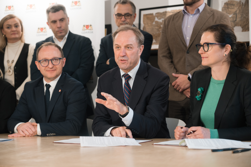 wicemarszałek województwa pomorskiego Marcin Skwierawski, marszałek województwa pomorskiego Mieczysław Struk oraz Prezydent Gdańska Aleksandra Dulkiewicz podpisują przy stole prezydialnym umowę. W tle widać kilka innych osób
