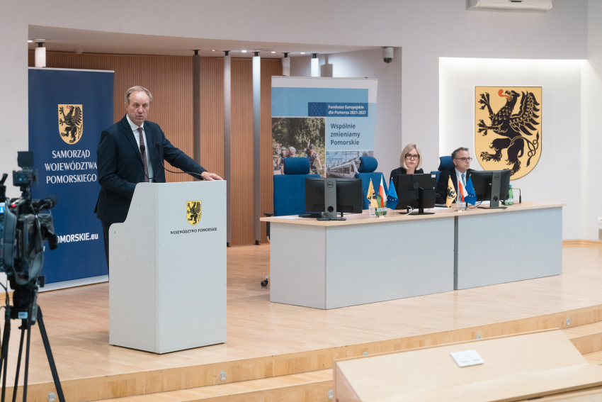 trzy osoby siedzą przy stole prezydialnym. Jedna osoba mówi przy mównicy