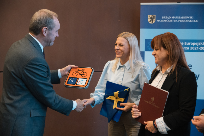 Marszałek Województwa Pomorskiego Mieczysław Struk wręcza znak szlaku rowerowego dwóm osobom, które trzymają flagę UE oraz podpisaną umowę o dofinansowanie