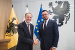 Marszałek Województwa Pomorskiego Mieczysław Struk oraz wójt gminy Arkadiusz Skorek pozują do zdjęcia i ściskają w geście powitalnym dłonie. W tle herb województwa pomorskiego, flaga województwa pomorskiego, flaga Polski oraz flaga UE