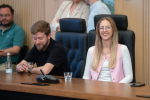 Dwie osoby siedzą i uśmiechają się podczas prezentacji. W tle widać inne trzy osoby