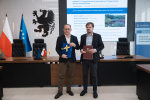 Wicemarszałek województwa pomorskiego oraz przedstawiciel Powiślańskiej Regionalna Agencji Zarządzania Energią pozują do uroczystego zdjęcia prezentując podpisane umowy o dofinansowanie oraz flagi UE