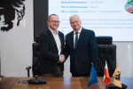 Wicemarszałek województwa pomorskiego oraz przedstawiciel lokalnych władz ściskają sobie dłonie przy stole prezydialnym