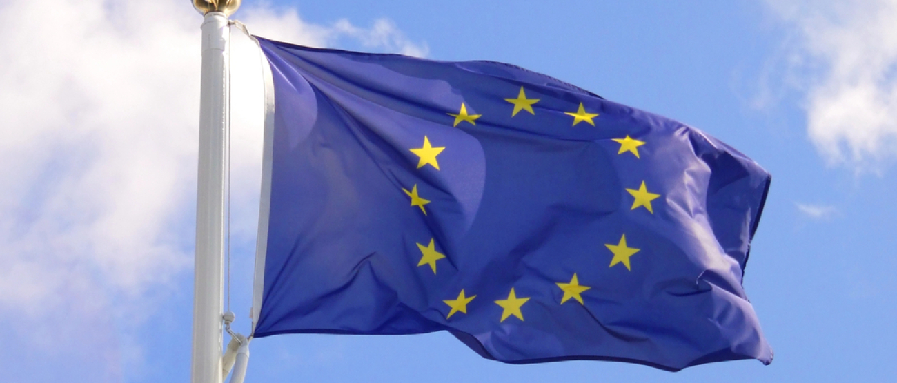 Flaga Unii Europejskiej w kształcie prostokąta powiewa na wietrze. Dwanaście żółtych gwiazd w kręgu na niebieskim tle.