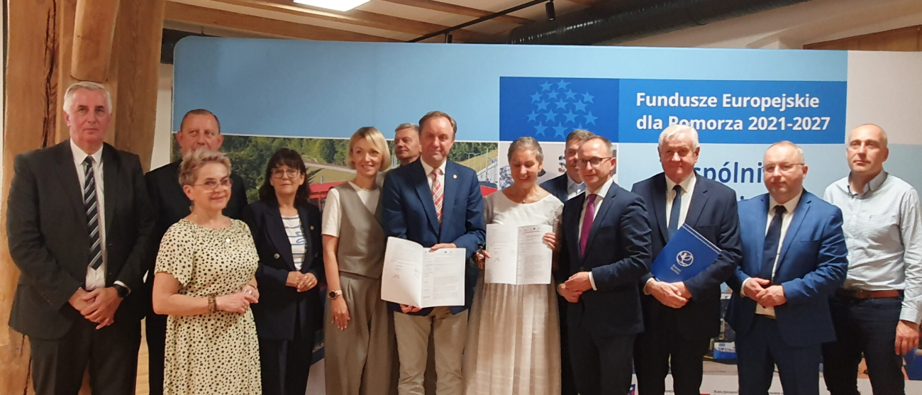 podpisanie umowy, 11 osób stoi na tle plakatu, marszałek trzyma podpisane porozumienie