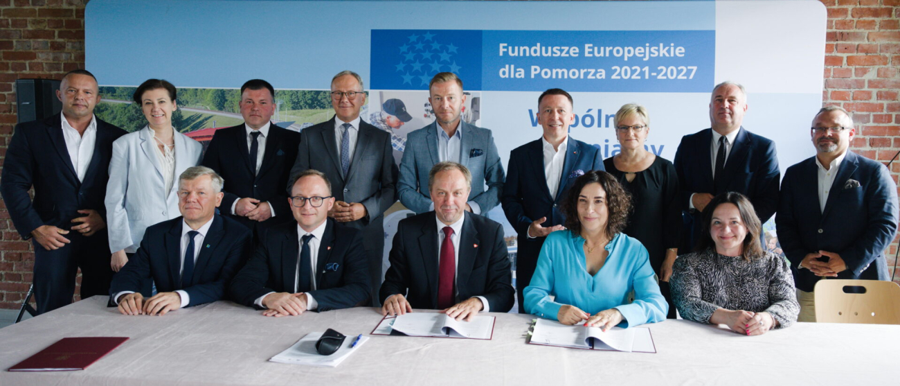 Grupa ludzi przy stole. Za nimi w drugim rzędzie stoja kolejne osoby. W tle ścianka Fundusze Europejskie dla Pomorza 2021-2027
