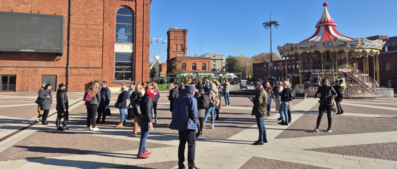Grupa ludzi stojąca na placu wyłożonym kostką brukową. W tle wyremontowane budynki z czerwonej cegły po dawnych fabrykach i duża karuzela