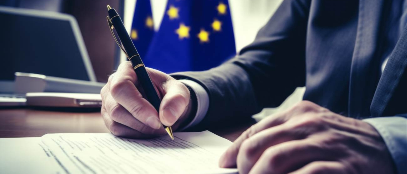 mężczyzna siedzący przy biurku, podpisuje dokumenty w tle flaga Unii Europejskiej 