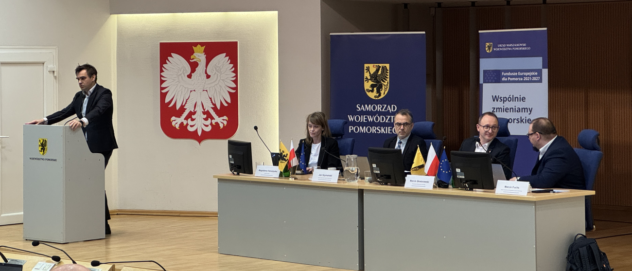 Osoby siedzą na sali. Cztery osoby siedzą w prezydium. Piąta osoba stoi przy mównicy.