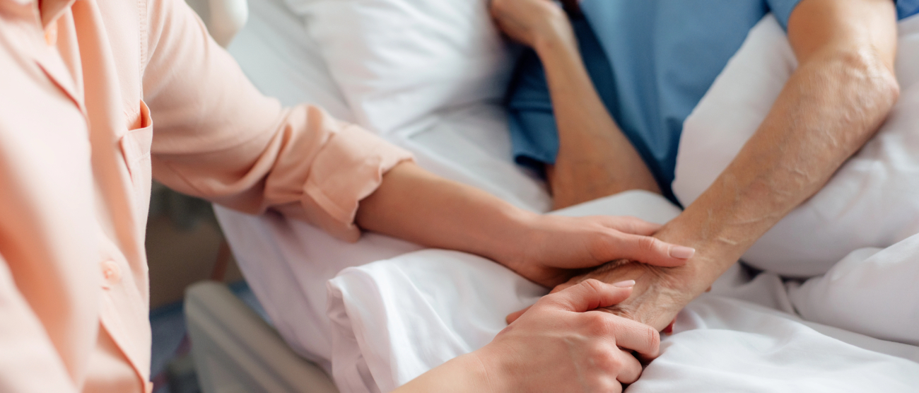 Kobieta leży łóżku. Druga trzyma jej dłoń