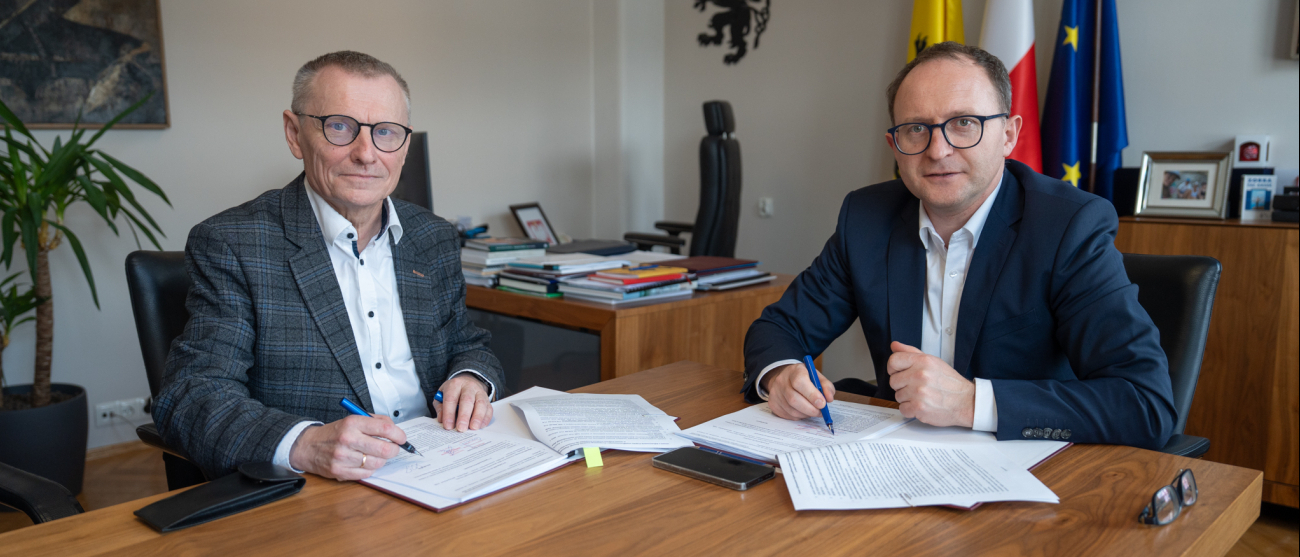 Dwie osoby podczas spotkania przy stole w biurze – dwie siedzą naprzeciw mężczyzny w garniturze, na stole leżą dokumenty, kubki z kawą i telefon.