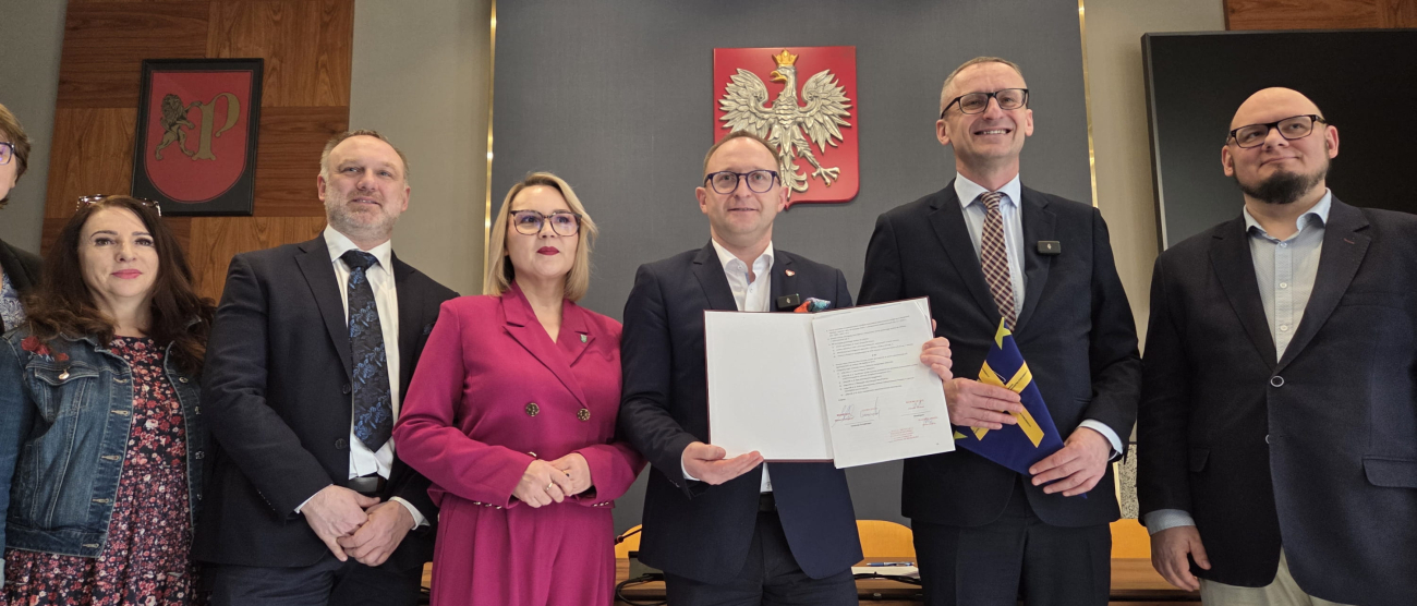 Grupa sześciu osób w strojach wizytowych stoi w rzędzie w oficjalnym pomieszczeniu. Centralna postać, mężczyzna w ciemnym garniturze i okularach, trzyma otwarty, podpisany dokument. Po jego prawej stronie stoi kobieta w wyrazistym, amarantowym garniturze. Na ścianie za nimi widnieje godło Polski (orzeł biały na czerwonej tarczy) oraz herb z lwem. W tle widać nowoczesne oświetlenie sufitowe oraz drewniane panele ścienne. Oficjalne podpisanie porozumienia lub uroczystość urzędową.