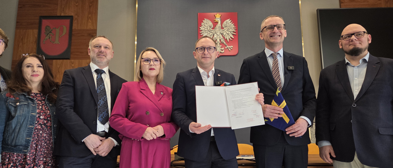 Grupa sześciu osób w strojach wizytowych stoi w rzędzie w oficjalnym pomieszczeniu. Centralna postać, mężczyzna w ciemnym garniturze i okularach, trzyma otwarty, podpisany dokument. Po jego prawej stronie stoi kobieta w wyrazistym, amarantowym garniturze. Na ścianie za nimi widnieje godło Polski (orzeł biały na czerwonej tarczy) oraz herb z lwem. W tle widać nowoczesne oświetlenie sufitowe oraz drewniane panele ścienne. Oficjalne podpisanie porozumienia lub uroczystość urzędową.