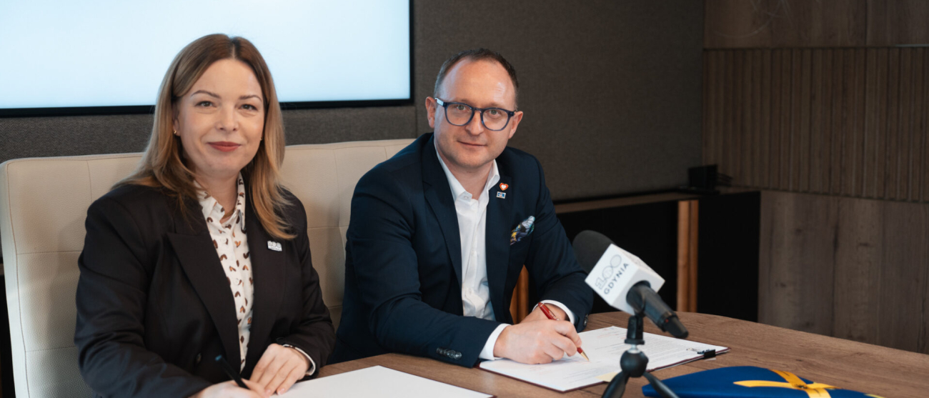 Dwie osoby w strojach biznesowych, kobieta i mężczyzna, siedzą przy drewnianym stole i podpisują dokumenty. Oboje uśmiechają się w stronę obiektywu. W tle widoczny jest duży ekran z napisem „GDYNIA 2026”, a na stole przed nimi stoi mikrofon z logotypem oraz flaga UE