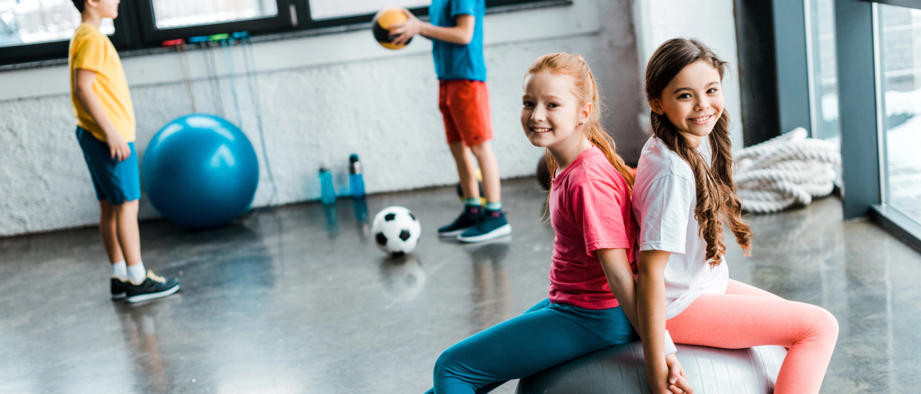 Dwie dziewczynki siedzą na dużej piłce treningowej. W tle dwóch chłopców rzuca do siebie piłkę. 