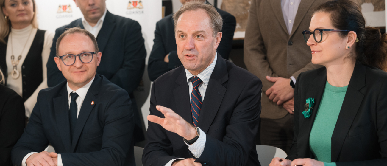 wicemarszałek województwa pomorskiego Marcin Skwierawski, marszałek województwa pomorskiego Mieczysław Struk oraz Prezydent Gdańska Aleksandra Dulkiewicz podpisują przy stole prezydialnym umowę. W tle widać kilka innych osób