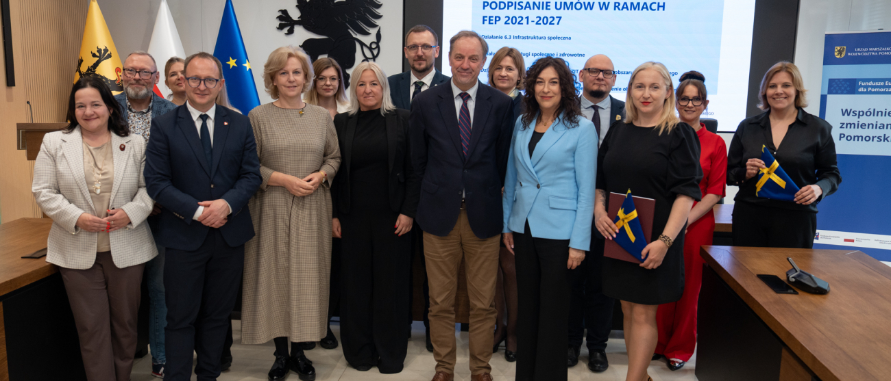 Szesnaście osób pozuje do uroczystego zdjęcia. Wśród zebranych znajduje się marszałek województwa pomorskiego Mieczysław Struk. Dwie osoby prezentują dokument i flagi Unii Europejskiej