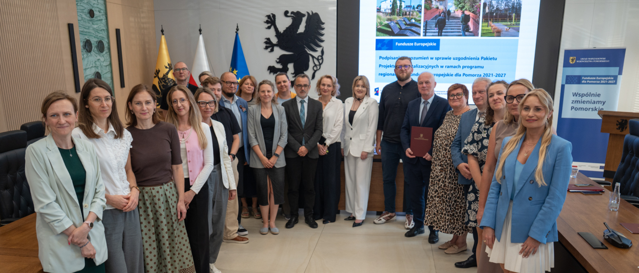Dwadzieścia dwie osoby pozują do uroczystego zbiorowego zdjęcia. W tle widać flagi województwa, Polski oraz Unii Europejskiej