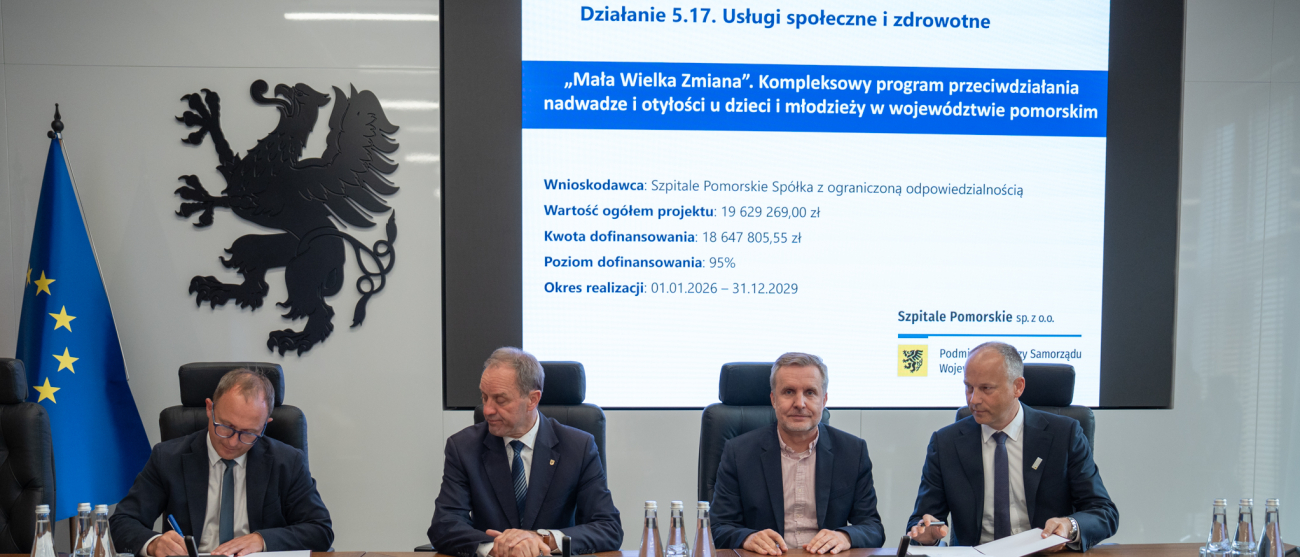 Cztery osoby podpisują umowę przy stole. W tle prezentacja. 