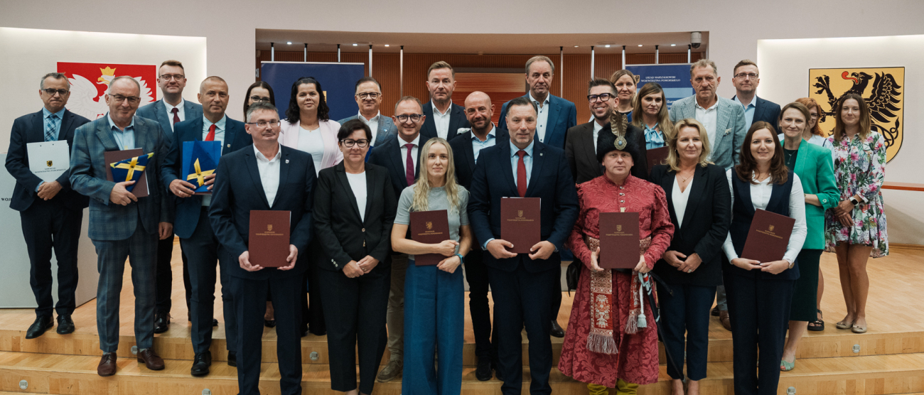 26 osób pozuje uroczyście do zdjęcia prezentując podpisane umowy oraz flagi UE