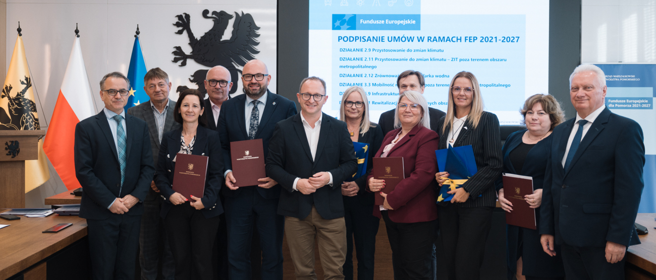 Dwanaście osób pozuje do uroczystego zdjęcia prezentując podpisane umowy o dofinansowanie oraz flagi UE - wśród nich wicemarszałek województwa pomorskiego oraz przedstawiciele lokalnych władz.