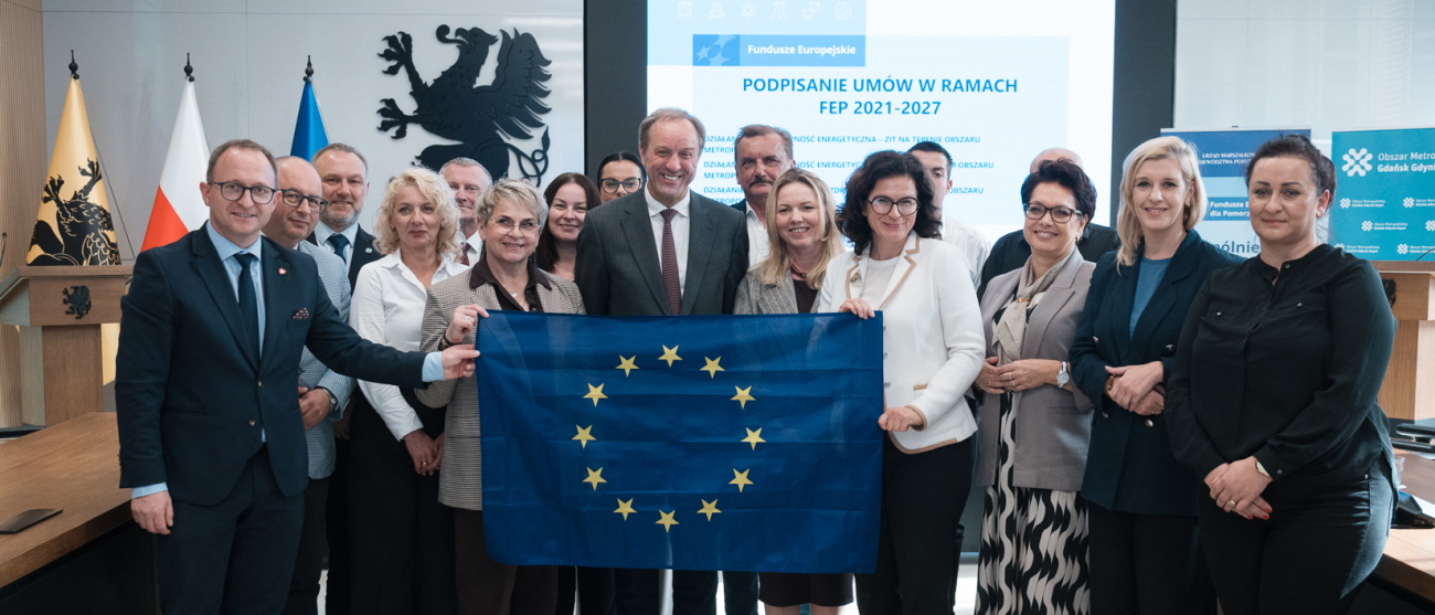 Siedemnaście osób pozuje do zdjęcia prezentując rozwiniętą flagę UE oraz podpisane umowy o dofinansowanie. Wśród tych osób jest marszałek woj. pomorskiego Mieczysław Struk i wicemarszałek Marcin Skwierawski