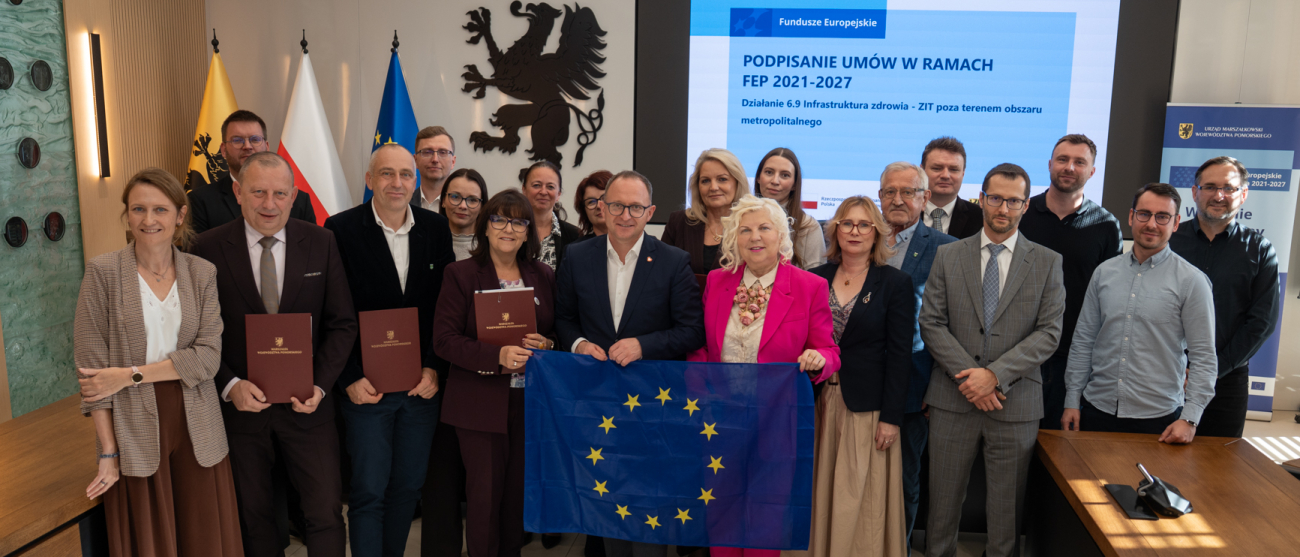 Dwadzieścia osób pozuje do uroczystego zdjęcia, prezentując podpisane umowy oraz rozwiniętą flagę Unii Europejskiej. Wśród zebranych znajduje się wicemarszałek województwa pomorskiego Marcin Skwierawski